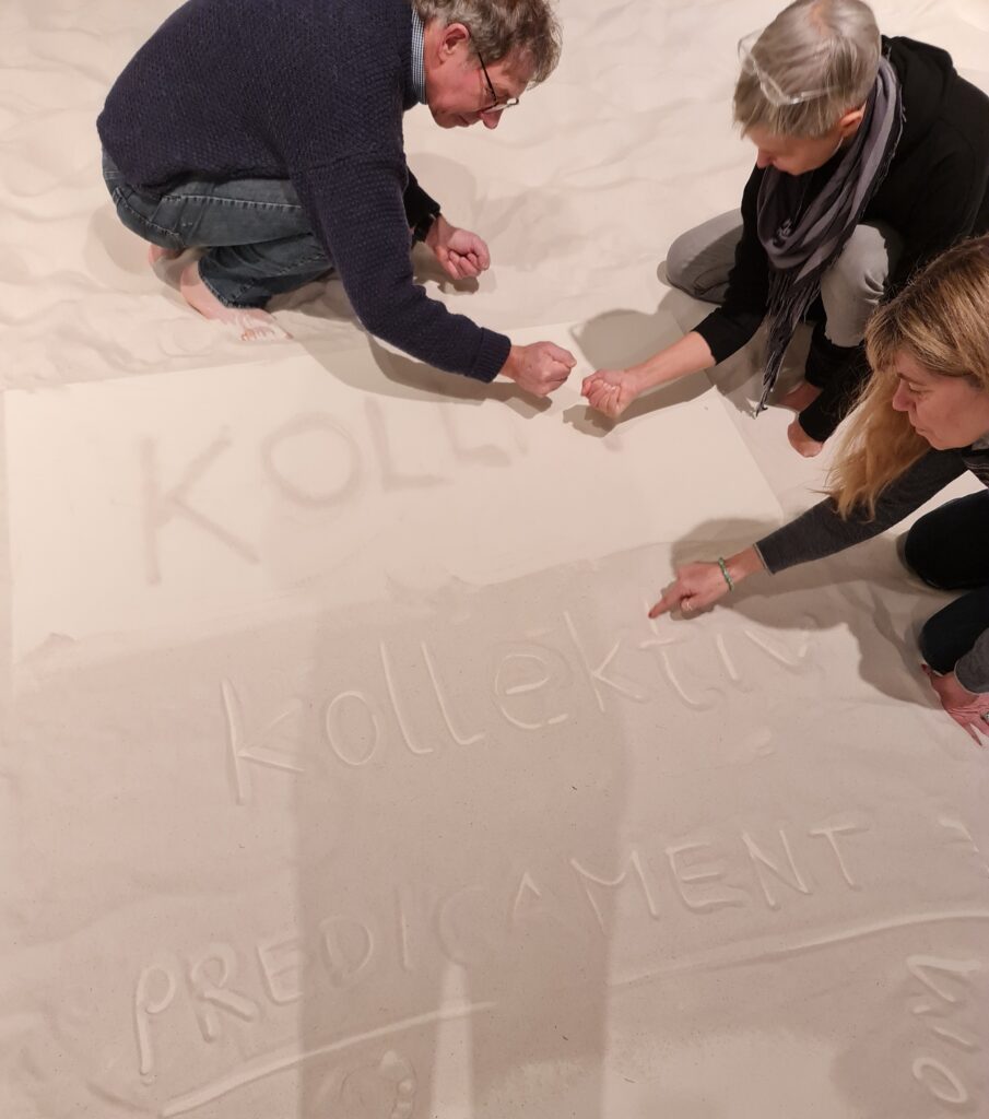 Drei Menschen schreiben in und mit Sand die Schriftzüge "Kollaps", "Kollektiv" und "Predicament".